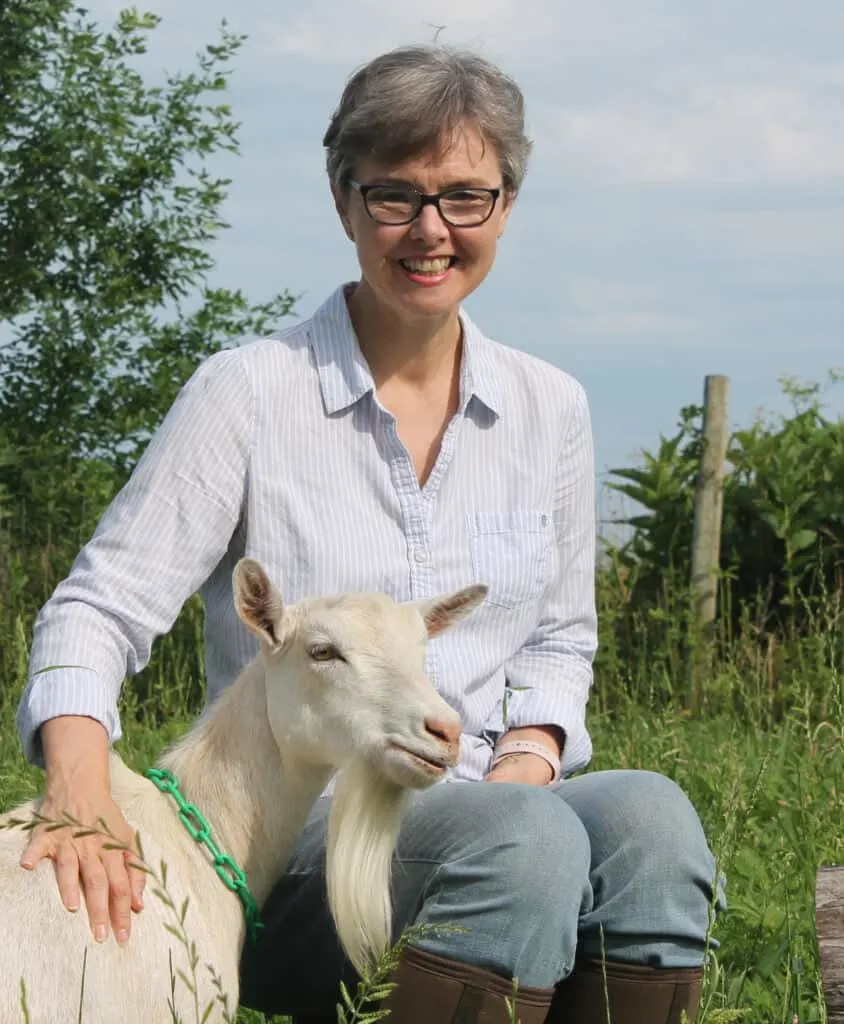 Deborah of Thrifty Homesteader smiling, sitting next to a white goat